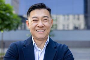 Portrait of a cheerful Asian businessman smiling confidently, shot outdoors with a blurred cityscape. photo