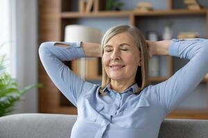 Portrait of satisfied female in blue blouse leaning on back of sofa and holding hands behind head with closed eyes. Smiling lady feeling comfortable while relaxing on furniture in stylish apartment. photo