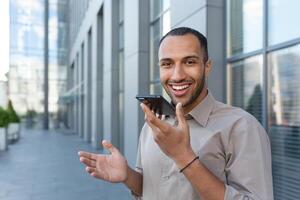 Outdoor portrait of young smiling African American man outside office building, man recording audio message smiling and looking at camera, businessman satisfied with work of AI voice assistant. photo