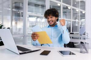 Businessman celebrating victory and success, receiving mail envelope with good news notification, hispanic reading document and happy holding hand up triumph gesture, man at workplace inside office. photo