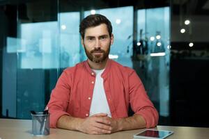 Online chat, remote business meeting, businessman in red shirt looking seriously thinking and focused into camera, listening to interlocutor, webcam view, inside office. photo