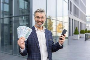 sorprendido empresario participación pila de dinero y teléfono en manos, contento con victorioso lotería, promoción a trabajar, mirando a cámara y sonriente. foto