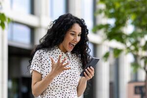 joven alegre mujer ganador recibido en línea notificación en teléfono, Hispano mujer con Rizado pelo celebrando éxito y triunfo caminando en ciudad cerca oficina edificio afuera. foto