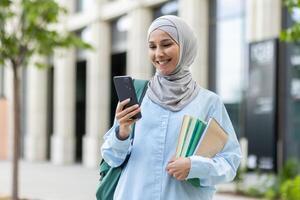 joven musulmán mujer en hijab caminando fuera de Universidad instalaciones, hembra estudiante sonriente contentamente utilizando aplicación en teléfono, mochila en espalda y libros en manos. foto