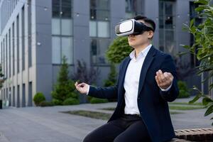 A young man in a business suit sits on a bench outside a company building in a lotus position, meditating in a virtual mask online. photo