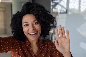 Beautiful business woman with curly hair wearing looking at smartphone camera, waving her hand in greeting gesture, talking with friends and colleagues, using call app photo