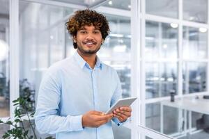 Portrait of young programmer with tablet computer inside office, hispanic man with curly hair smiling and looking at camera, man at workplace testing online applications standing near window. photo