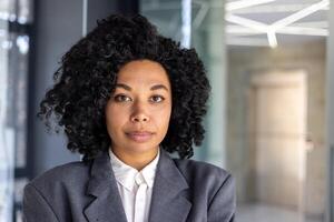 Close up portrait of serious confident business woman, african american woman with curly hair and in business suit looking at camera, female worker inside office at workplace. photo