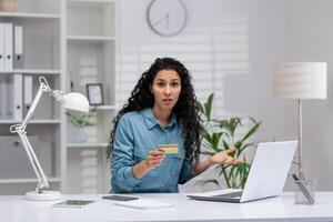 Professional Hispanic woman facing a problem while working on her laptop from the home office, confusion evident in her posture. photo