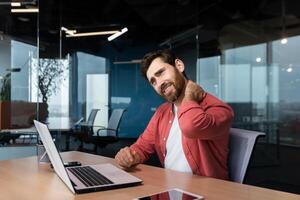 Overtired man in red shirt works inside office with laptop, businessman has severe neck pain, massages muscle with hand from sedentary work. photo