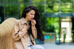 Worried woman coughing outdoors, feeling unwell. Health, sickness, public concern concepts in urban setting. photo