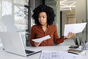 triste decepcionado mujer de negocios dentro oficina a lugar de trabajo mirando a documentos documentos financiero informes, africano americano hembra trabajador en papel trabajo Deprimido infeliz con financiero logros foto