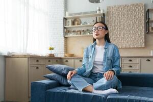 yoga a hogar. un joven asiático mujer en lentes se sienta calma y relajado a hogar en el sofá en el loto posición, cerrado su ojos, meditando foto