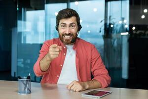 retrato de un joven hombre en un auriculares quien es emocionado hablando a el cámara, ondulación su manos expresivamente. se sienta en el oficina a el mesa, comunica vía llamar, consultas, comentarista. foto