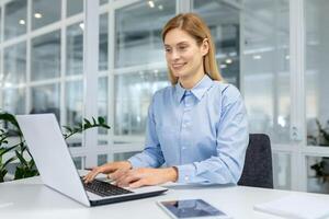 Smiling secretary keeping hands on keyboard of contemporary laptop while sitting on dark office chair. Adult lady using modern technology for developing possibilities of programming on distance. photo