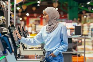 un mujer comprador en un supermercado en un hijab paga para bienes a un autoservicio verificar, conveniente Servicio para clientes foto