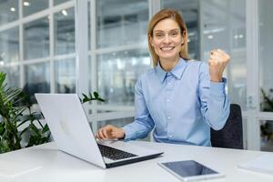 Successful woman looking at camera with toothy smile and making winner gesture in front of table with pc. Caucasian female in formal clothes clenching fist with pleasure from career achievement. photo