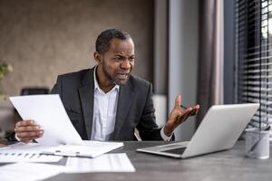 A professional African American businessman appears distressed while working with papers and a laptop in a modern office setting. photo