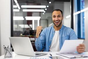 Successful Indian businessman fist-pumping in celebration at his modern office workspace, feeling excited and happy. photo