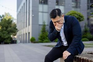 Asian young man in a suit sits on a bench in the street with his hand covering his mouth, feels severe nausea and urge to vomit. photo