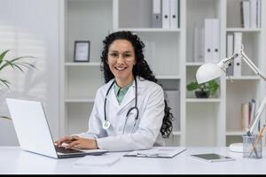 A cheerful Latino female doctor in a white coat smiles as she works on her laptop in a modern office setting. photo