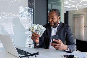 contento afroamericano hombre sentado a el escritorio en el oficina, participación un móvil teléfono en su mano y mirando con entusiasmo a un ventilador de efectivo dinero. foto
