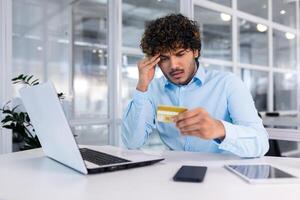 Problems with the account, bankruptcy, etc. A young hispanic man is sitting at the desk in the office, looking worriedly at the credit card, holding his head. photo