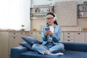 un joven asiático estudiante niña es sentado en el sofá a hogar en el loto posición. él escucha a un podcast, un audio libro, estudios en blanco auriculares con un teléfono en su manos. foto