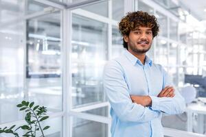 Young successful hispanic businessman in the middle of the office, portrait of happy and satisfied office worker, man in shirt and arms crossed smiling and looking at camera, businessman at work. photo