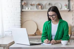 un joven mujer periodista, escritor, blogger trabajos remotamente desde hogar utilizando un ordenador portátil en línea y notas foto