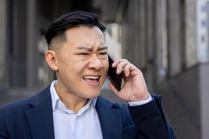 Annoyed Asian adult male in a business suit on a call outdoors, showing signs of frustration and urgency. photo