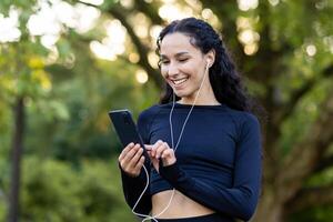 alegre activo joven mujer con auriculares sonriente mientras participación un teléfono, disfrutando un descanso en un lozano parque. foto