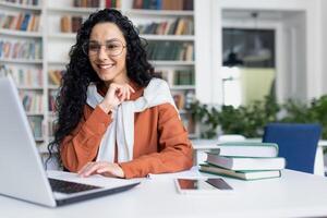 hembra estudiante con ordenador portátil leyendo en línea curso, latín americano mujer sonriente y satisfecho con independiente en línea aprendizaje sentado dentro Universidad instalaciones en biblioteca. foto