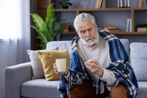 mayor caucásico hombre envuelto en un frazada, sentado en un sofá y comprobación su fiebre con un termómetro. él sostiene un taza, mirando preocupado y indispuesto en su vivo habitación. foto