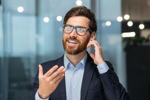 de cerca foto. un joven hombre en el oficina y hablando mediante un auriculares mediante su manos gratis. él es participación un auricular con su mano, gesticulando con su mano, sonriente. foto