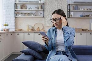 joven hermosa asiático trastornado mujer sentado a hogar en vivo habitación en sofá vistiendo lentes foto