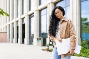 confidente joven mujer de negocios con ordenador portátil y cuaderno en pie en frente de un oficina edificio, exudando profesionalismo y ambición. foto