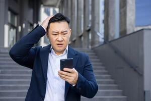 A confused Asian businessman reads a text on his phone, frowning with hand on head, standing on stairs outside. photo