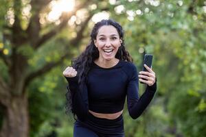 joven hermosa Hispano mujer contento sonriente y mirando a cámara, mujer en Mañana correr y activo ejercicio en parque, contento con rutina de ejercicio resultado, utilizando aplicación en teléfono. foto