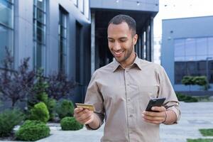 joven empresario empresario hace en línea compras, usos un teléfono inteligente y un banco crédito tarjeta, un hombre fuera de un oficina edificio camina alrededor el ciudad durante un descanso a trabajo foto