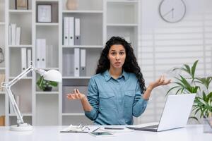 Perplexed Hispanic woman at her home office desk expressing confusion while using a laptop, possibly during a business issue. photo