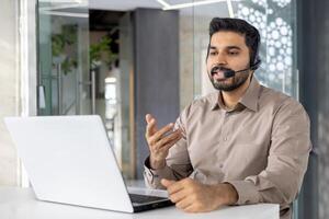 un joven profesional hombre atractivo en cliente Servicio utilizando un auriculares en un bien iluminado moderno oficina. él gestos mientras discurso, enfatizando activo comunicación. foto