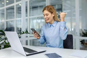 A professional woman in a modern office space celebrates success with a fist pump while holding her smartphone. photo