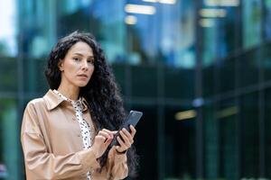 Serious mature woman boss outside office building looking thoughtfully at camera business woman holding phone in hands using app on smartphone, Hispanic woman with curly hair browsing internet pages photo