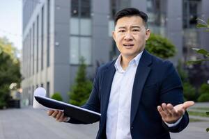 Close-up portrait of a young Asian man in a business suit standing outside an office building, holding documents and looking worriedly at the camera. photo