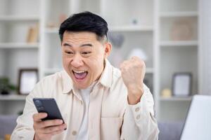 The winner is sitting at the table at home, the man is happy to receive the online notification of the victory and success on the phone, the Asian man is holding his hand up and holding smartphone. photo