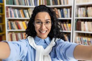 Confident Hispanic student captures a selfie moment surrounded by books in a library setting, portraying knowledge and education. photo