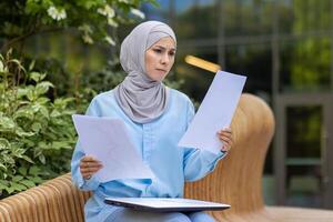 A focused professional woman in a hijab examines papers, sitting on an outdoor bench in a tranquil setting. photo