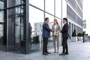 Meeting of three colleagues from outside the office building, experienced and mature IT specialists, greeting and shaking hands, business persons in business suits, diverse group of people photo