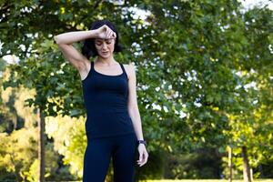 joven mujer trotar y haciendo Deportes en el parque, en pie en ropa de deporte y participación su cabeza con su mano, sensación un dolor de cabeza y limpiando sudor desde su frente cuando ella es cansado. foto